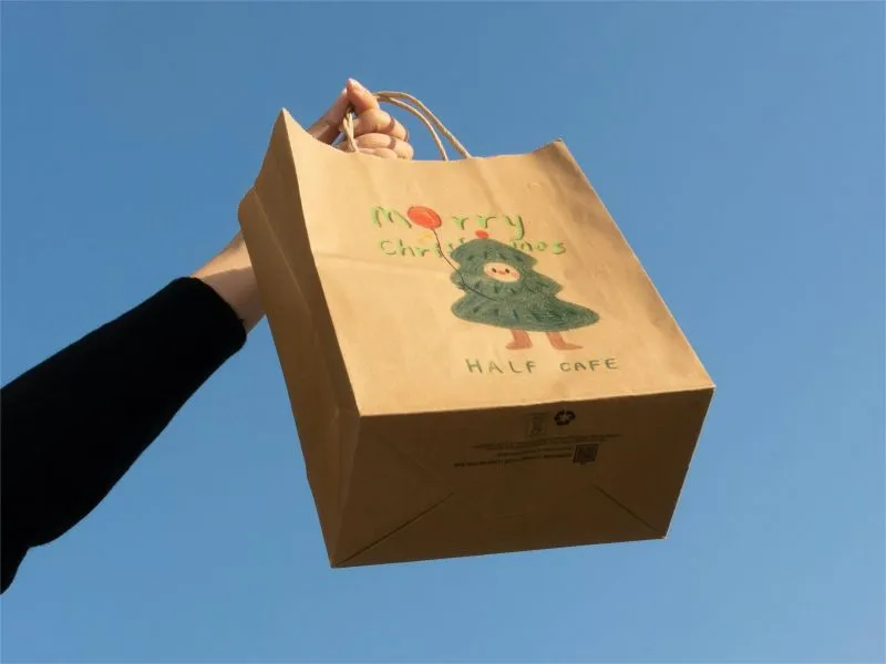 A hand holding a brown paper bag with a Christmas tree design and the text "Merry Christmas" and "Half Cafe" against a clear blue sky.