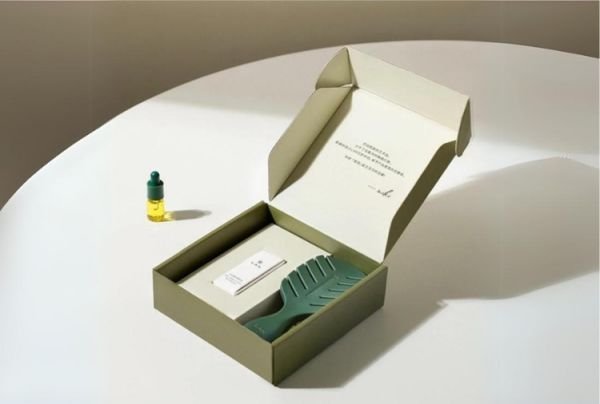 Open custom mailer box with an olive green exterior and beige interior, containing a small bottle, a product box, and a leaf-shaped insert, displayed on a white surface.