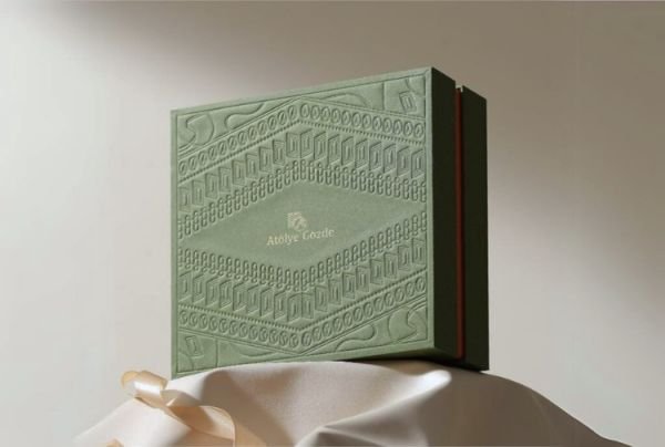 Elegant green custom rigid box with intricate embossed patterns and the text 'Above Grade' displayed in the center, set against a light fabric backdrop.